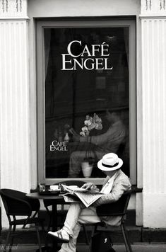 a man sitting at a table in front of a cafe reading a book while wearing a hat