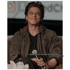 a man sitting in a chair holding a microphone and smiling at the camera while wearing a brown leather jacket