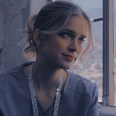 a woman in scrubs looking out a window at the city below her, with lights on