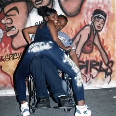 a woman sitting on top of a man in a wheel chair next to a mural