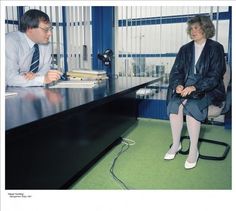 two people are sitting at a desk in an office setting, one is talking to the other