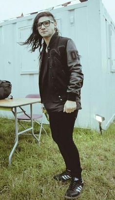 a man with long hair and glasses standing in the grass near a table, next to a portable toilet