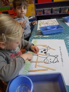 two young children sitting at a table making popsicle stick art work for the camera