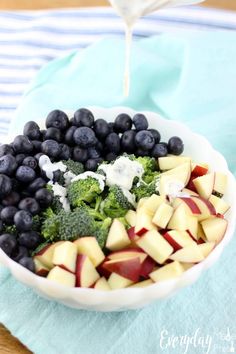 blueberries, broccoli and apples are in a bowl with dressing being poured over them