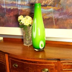 a green vase sitting on top of a wooden dresser next to a white flower arrangement