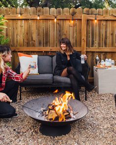 two women sitting around a fire pit with drinks in their hands and food on the table