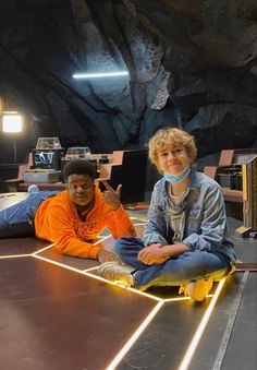 two boys sitting on the ground in front of a cave