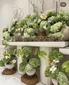 several white vases filled with flowers and greenery on top of a wooden shelf