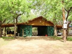 a small green building surrounded by trees in the middle of a dirt field with cars parked nearby