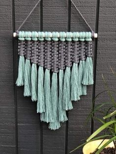 a wall hanging made out of yarn and tassels on a black fence next to a potted plant