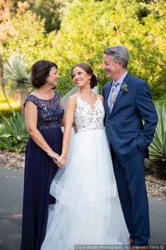 two women and a man standing next to each other in front of trees with bushes