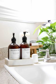 two bottles of hand soap sitting on top of a sink next to a green plant