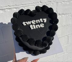 a black heart shaped cake with the words twenty nine on it, held up against a white brick wall