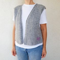 a woman standing in front of a white wall wearing a gray sweater vest and jeans