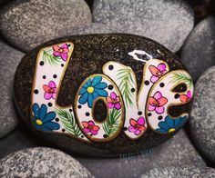 a rock with the word love painted on it surrounded by flowers and leaves in front of some rocks