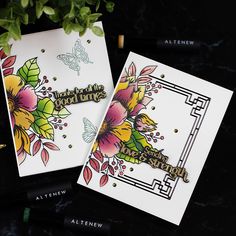 two cards with flowers on them sitting next to some pens and pencils in front of a potted plant