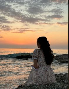 a woman sitting on top of a rocky beach next to the ocean at sunset or dawn