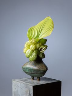 a green vase with flowers in it on top of a wooden block against a gray background