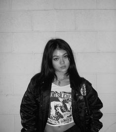 a woman standing in front of a white brick wall wearing a black jacket and t - shirt