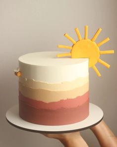 a person holding up a cake with the sun on top and pink, yellow, and white frosting