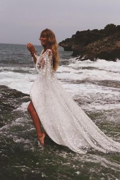 a woman in a white dress is standing on the rocks near the ocean and waves