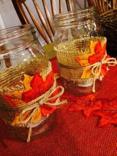 three mason jars with fall leaves in them sitting on a red table cloth next to wicker baskets