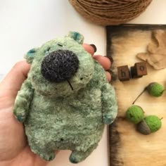 a hand holding a green teddy bear next to some balls of string and yarn on a table