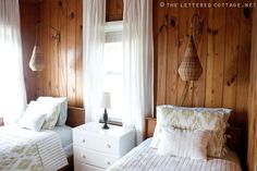 two beds in a room with wood paneling and white curtains on the windowsill