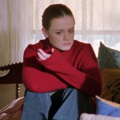 a woman sitting on top of a bed in a red sweater and blue jean pants