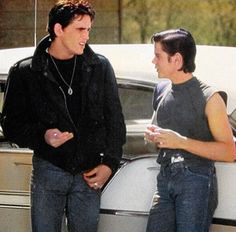 two young men standing next to each other near a parked car, talking and laughing