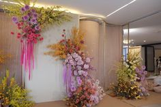 there are many different types of flowers on display in front of a wall that is decorated with sheer curtains