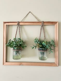 two mason jars with plants hanging from them on a string and wood frame hung on the wall