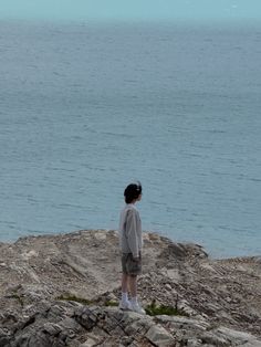 a young boy standing on top of a rocky hill next to the ocean with a kite flying in the sky