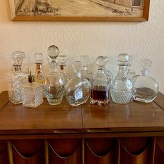 an assortment of glass bottles and decanters on a wooden table in front of a painting