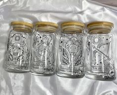 four glass jars with drawings on them sitting on a white cloth lined up in a row