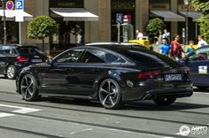 the black car is driving down the street in front of other cars and onlookers