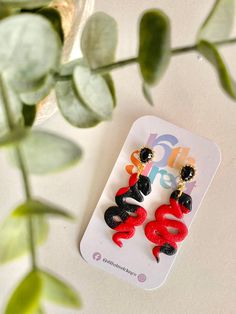 two red and black earrings sitting on top of a white table next to a plant