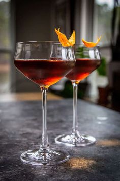 two glasses filled with red wine and orange peels on a counter top next to each other