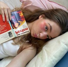 a woman laying in bed with a book on her head and eye makeup painted pink