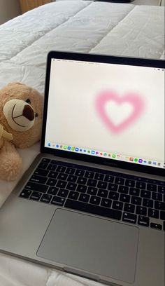 a teddy bear sitting next to a laptop on a bed with a heart drawn on the screen