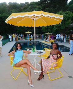 two women sitting at a table with an umbrella over them in front of a pool