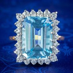 An imposing Art Deco style cocktail ring built around a large emerald cut blue topaz, weighing an impressive 9 carats (approx.) It has a deep ocean blue hue, similar to an aquamarine and is framed by twinkling brilliant cut cubic zirconia set around the border. The rectangular gallery consists of slender claws that wrap around each stone to form a fabulous, tiered bed for the stones to rest on. It's all silver and the band has been gilded in 18ct yellow gold. It's a fantastic statement piece and Blue Octagon Ring With Halo Setting, Blue Octagon Halo Setting Rings, Octagon Blue Rings With Halo Setting, Blue Octagon Rings With Halo Setting, Blue Aquamarine Emerald-cut Diamond Ring, Formal Blue Emerald Ring With Halo Setting, Blue Emerald-cut Aquamarine Diamond Ring, Blue Topaz Ring With Emerald-cut Halo Setting, Blue Emerald-cut Topaz Ring With Halo Setting