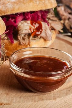 a bowl of bbq sauce next to a sandwich on a cutting board with meat in the background