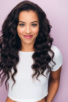 a woman with long curly hair smiling at the camera, wearing a white crop top