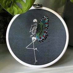 a cross - stitched skeleton with a butterfly on it's back sits in front of a potted plant