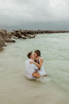 Young newly engaged couple kiss intimately, with her sitting in his lap on water's edge of surf at sunrise in PCB Florida.  Photo taken by couples photographer Brittney Stanley of Be Seen Photos