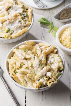 two white bowls filled with pasta and chicken casserole next to silver spoons