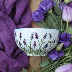 a white bowl with purple flowers sitting on top of a wooden table next to purple fabric