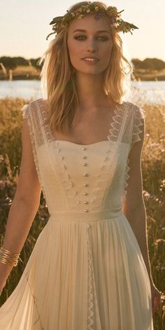 a woman wearing a white dress standing in a field with flowers on her head and hands behind her back