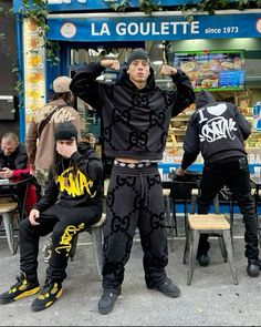 three men in black and yellow clothing sitting on chairs outside a store front with their hands up